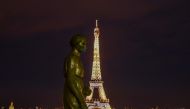 A picture taken on May 2, 2020 shows a bronze statue wearing a face mask emmulating the actions of many citizens to protecting themselves against the novel coronavirus, COVID-19 at the Parvis des Droits de l'Homme, with the Eiffel Tower enlightened in bac