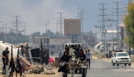 A man and woman ride in a donkey-drawn cart carrying a water cistern while behind smoke billows from Israeli bombardment, along Salah al-din road in Deir el-Balah in the central Gaza Strip on July 21, 2025. (Photo by Eyad BABA / AFP)