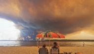 Beach goers sit on the beach of Moutchic in Lacanau, southwestern France on July 24, 2026, as clouds of smoke rise into the sky from a wildfire. Photo by Maximilien Lamy / AFP