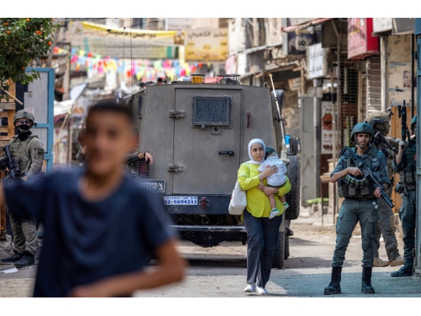 A Palestinian woman carrying a child walks past members of the Israeli occupation army and armoured vehicles during an Israeli military operation at the Balata camp for Palestinian refugees, east of Nablus in the occupied West Bank, on August 31, 2026. Photo by JAAFAR ASHTIYEH / AFP