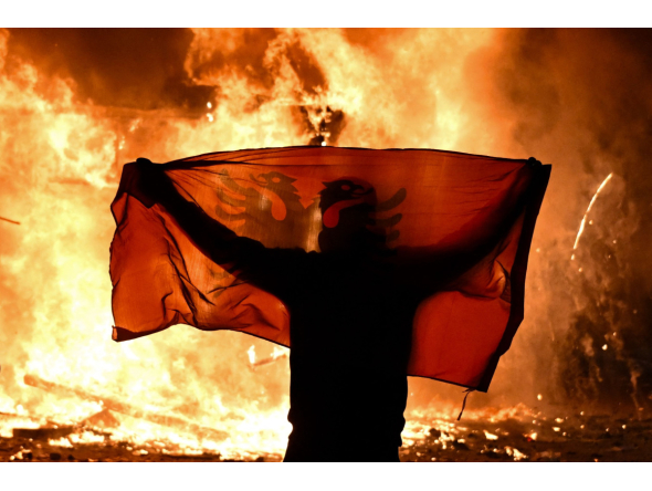 A protester holds an Albanian National flag in front of a burning kiosk during an anti-government demonstration in central Tirana on October 15, 2026, as protests continued for a third consecutive day over university conditions and demands for the resignation of Albanian Prime Minister. Photo by Armend NIMANI / AFP