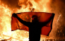 A protester holds an Albanian National flag in front of a burning kiosk during an anti-government demonstration in central Tirana on October 15, 2026, as protests continued for a third consecutive day over university conditions and demands for the resignation of Albanian Prime Minister. Photo by Armend NIMANI / AFP