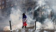 A protester, wrapped in a flag of France, stands in the smoke at Place de l'Opera during ongoing protest action, in Lille, northern France, on October 17, 2026. (Photo by Sameer Al-Doumy / AFP)