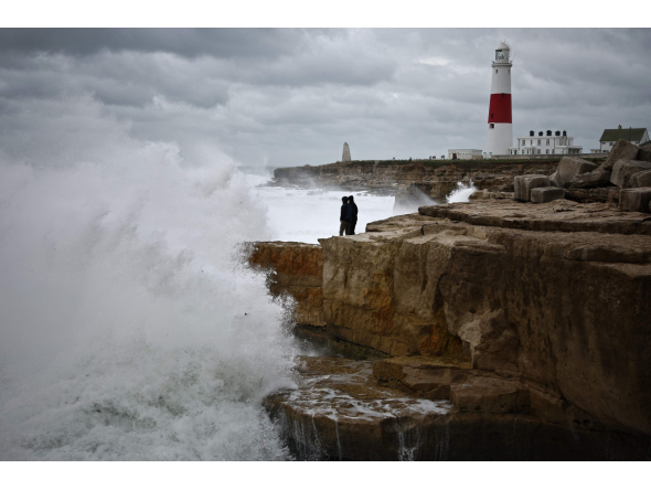 Members of the public stand on rocks as waves break near Portland Bill Lighthouse in Portland, southern England on October 23, 2026, as Storm Austen brings strong winds and heavy rain across much of southern England. (Photo by Henry Nicholls / AFP)