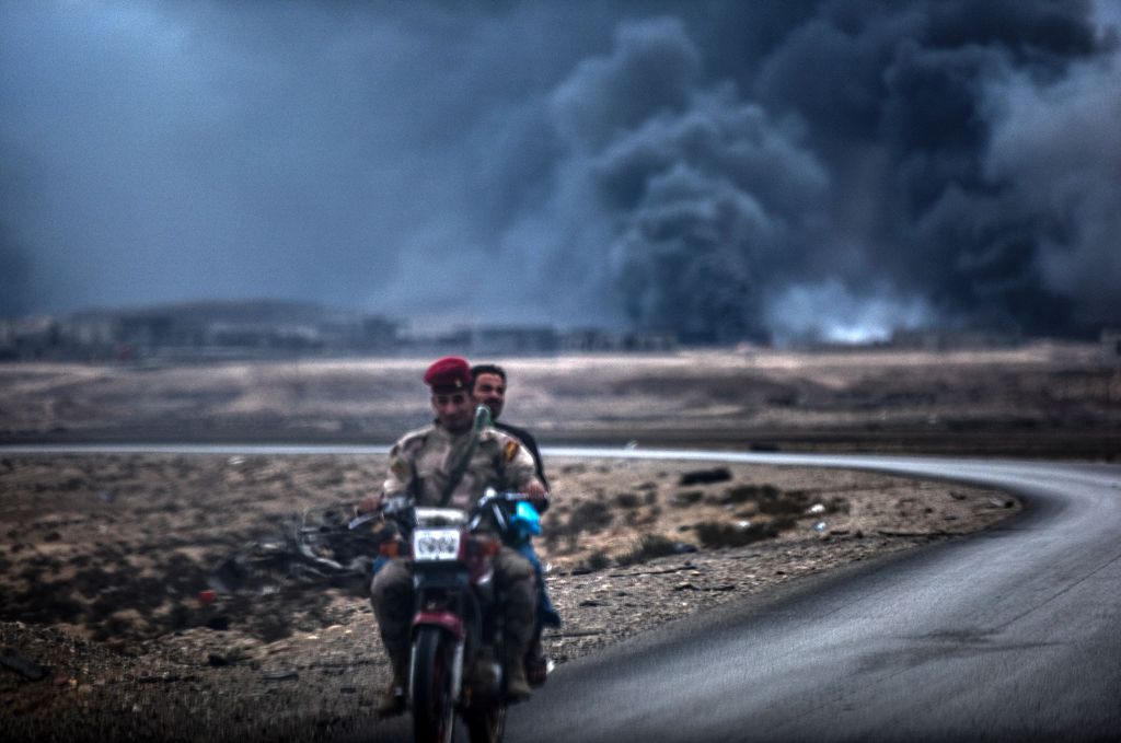 An Iraqi soldier and a civilian ride a motorbike as smoke rises behind them, on the road between Qayyarah and Mosul on October 28, 2016.  AFP / BULENT KILIC
