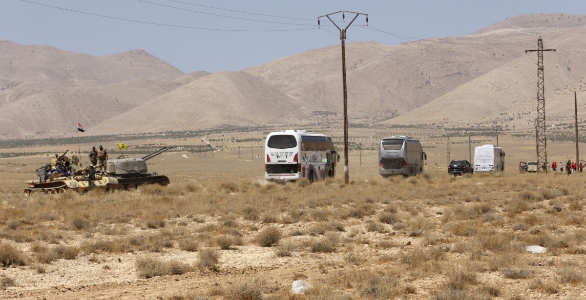 Syrian forces members stand on a tank next to vehicles waiting to transport Islamic State (IS) group members in the Qara area in Syria's Qalamoun region on August 28, 2017 as part of a deal between Hezbollah and IS fighters where the jihadists would leave