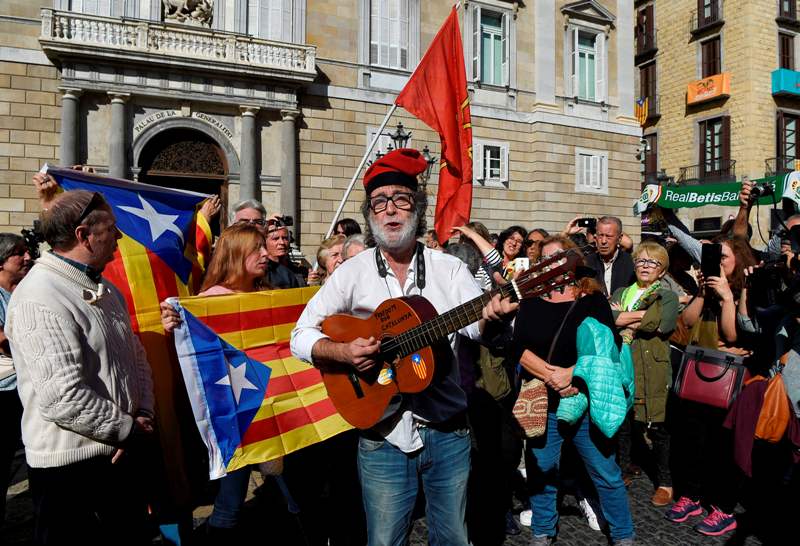 A man sporting a Catalan typical 'barretina' hat plays guitar as other people wave Catalan flags in front of the 'Generalitat' palace (Catalan government headquarters) in Barcelona on October 30, 2017.  AFP / LLUIS GENE
