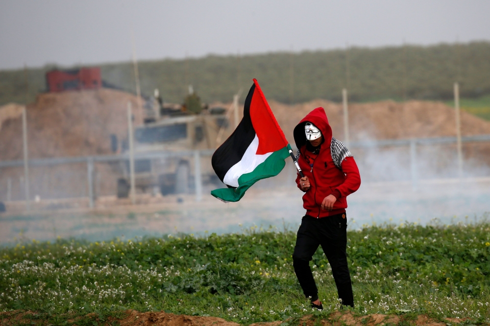 A Palestinian protester wearing a Guy Fawkes mask carries the national flag during clashes with Israeli forces following a demonstration near the fence along the border with Israel, east of Gaza City, on February 15, 2019. AFP / Said Khatib 
