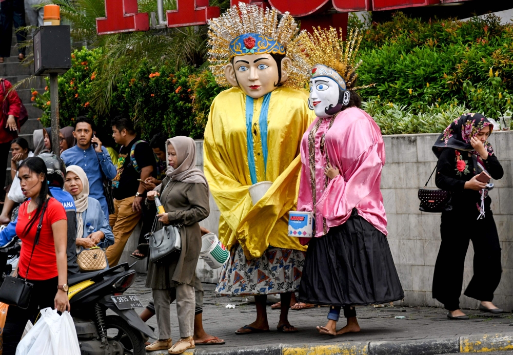 This picture taken on April 26, 2019 shows people walking past Ondel-ondel effigies worn by children busking for money on a street in Jakarta.  AFP / Goh Chai Hin 