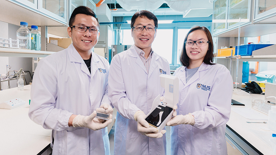 The NUS research team behind the novel algae detection device is led by Assistant Professor Bae Sung Woo (centre) who is holding the smartphone platform. With him are two team members: Mr Thio Si Kuan (left) who is holding the microfluidic chip, and Miss 