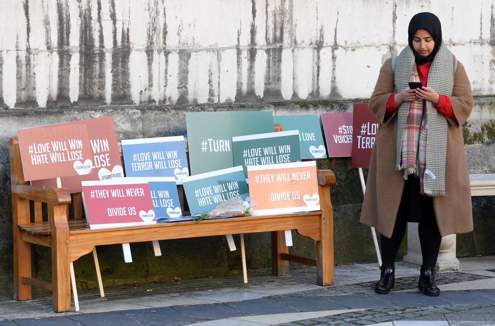 A woman checks her mobile phone next to signs with slogans during a vigil for victims of a fatal attack on London Bridge in London, Britain December 2, 2019. Reuters/Toby Melville
 