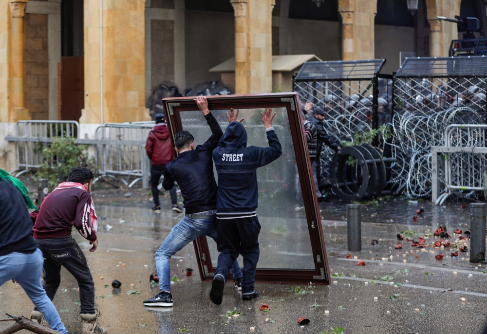 Anti-government protesters use a window as a makeshift shield as they confront security forces from behind a barbed-wire and metal fence barrier during clashes in the central downtown district of the Lebanese capital Beirut near the parliament headquarter