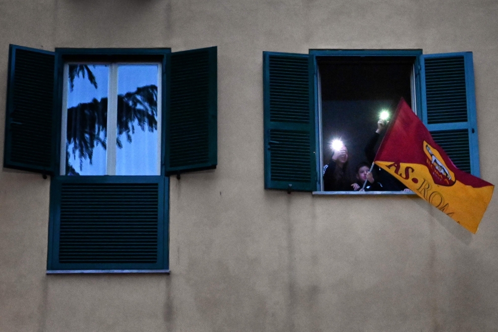 People wave an AS Roma flag at their windows as they listen to artists performing in the courtyard of a popular apartment building for the show Sotto lo Stesso Cielo tour (Under the Same Sky tour) in San Basilio suburbs of Rome on April 18, 2020, during t
