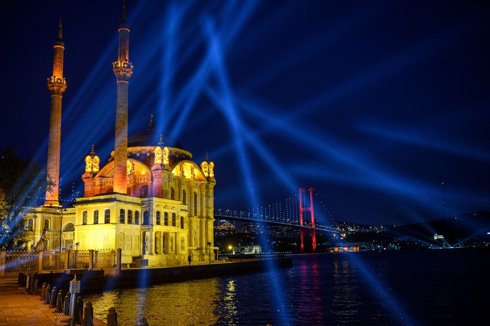 TOPSHOT - A picture taken on May 19, 2020 shows the Bosphorus bridge and the Ortakoy mosque as a light shine over the Bosphorus to mark the Youth and Sports Day in Istanbul during a four-day curfew aimed at curbing the spread of the COVID-19 pandemic, cau