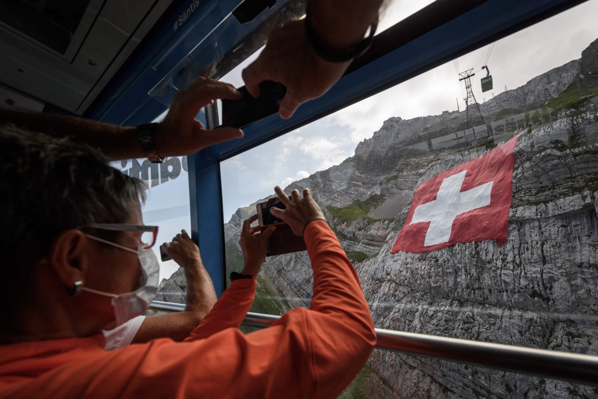 Tourists wearing a face mask take pictures of a huge 6400 meters square Swiss flag hanging on mountain Saentis on eve of Swiss national day on July 31, 2020 near Schw?galp, eastern Switzerland. Many Swiss stay in their country for holidays due to travel r