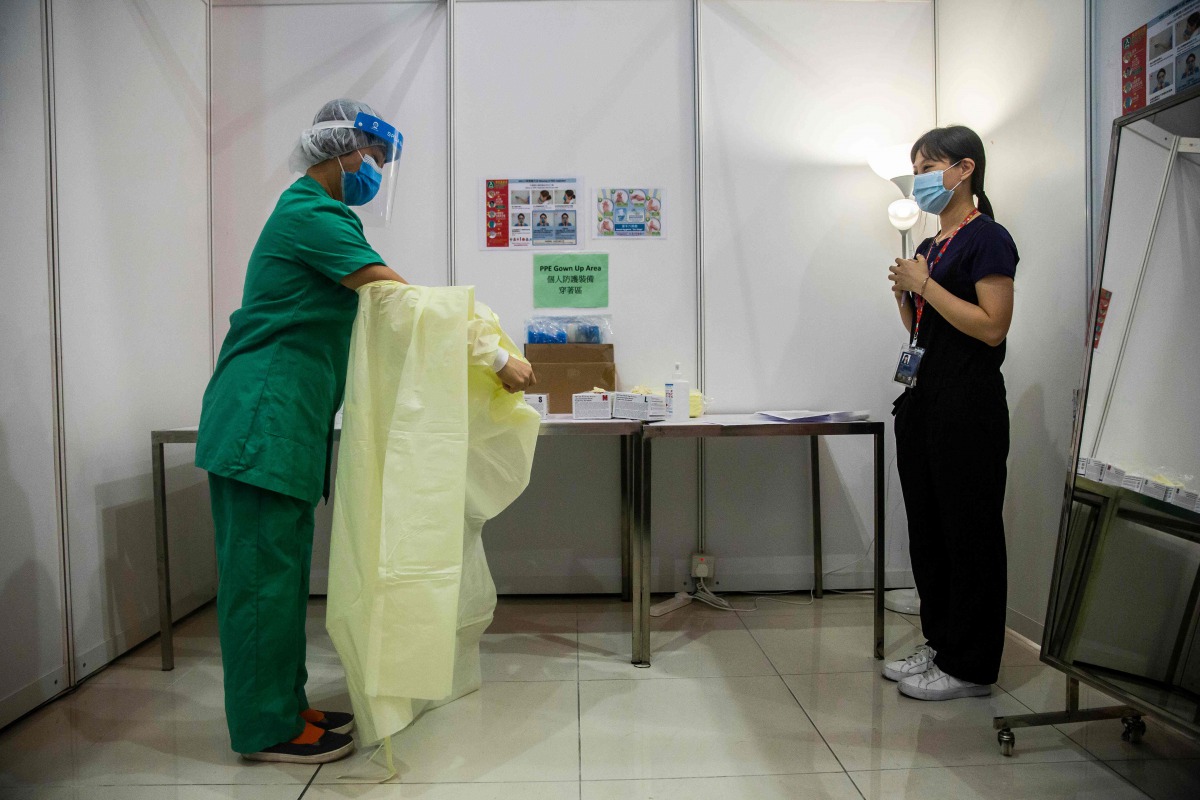 A medical worker (L) demonstrates removing her gown while being supervised in an area where protective gear is put on and taken off at a temporary field hospital set up at Asia World Expo in Hong Kong on August 1, 2020. / AFP / ISAAC LAWRENCE