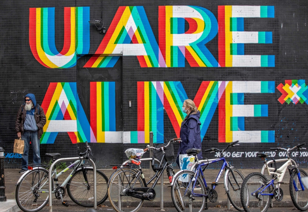 A pedestrian wearing a face mask or covering due to the COVID-19 pandemic, walks past a mural reading 