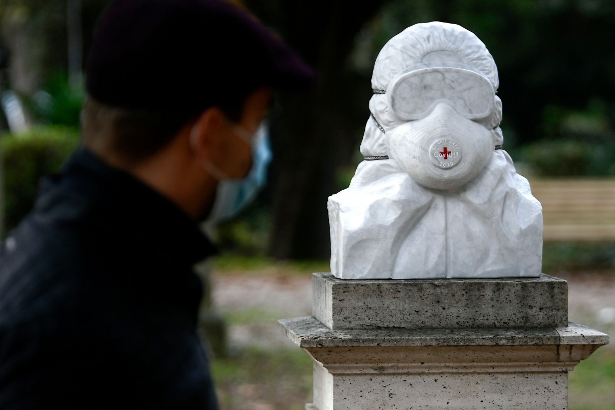 A man wearing a face mask walks by a marble bust dedicated to doctors, nurses and health workers untitled 