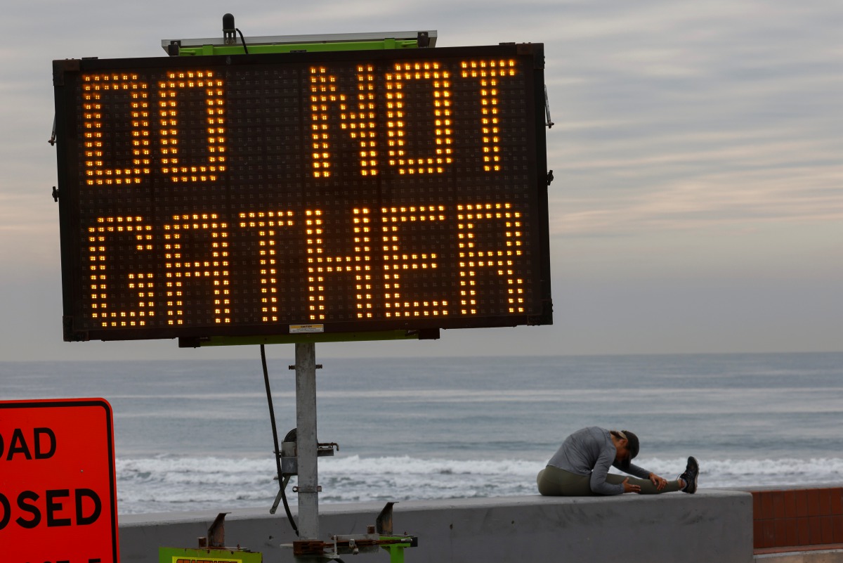 A woman stretches near a warning sign on the beach as new stay-at-home orders begin in Southern California during the outbreak of the coronavirus disease (COVID-19) in Oceanside, California, U.S., December 7, 2020. REUTERS/Mike Blake
