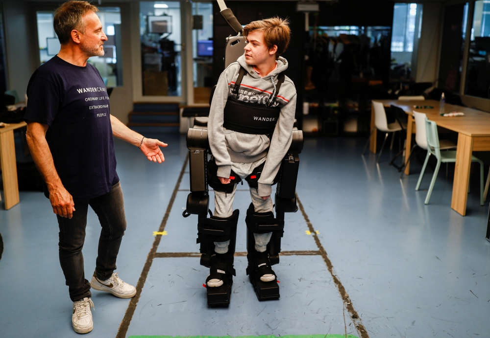 :Jean-Louis Constanza, Chief Business and Clinical Officer at French company Wandercraft helps his 16-year-old son Oscar using a robot exoskeleton at the company headquarters in Paris, France July 9, 2021. Picture taken July 9, 2021. REUTERS/Christian Har