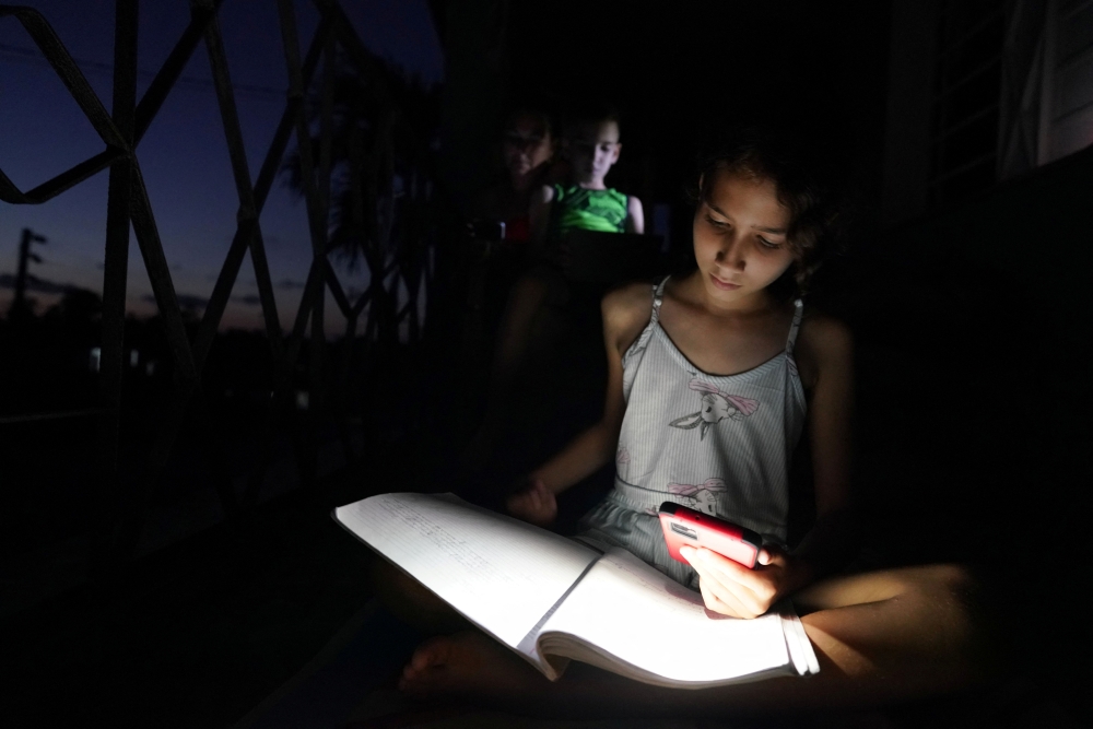 Emily Villega, 10, does her homework using the light of a cell phone at the balcony of her home during a blackout in Guanajay, Cuba June 17, 2022. Reuters/Alexandre Meneghini