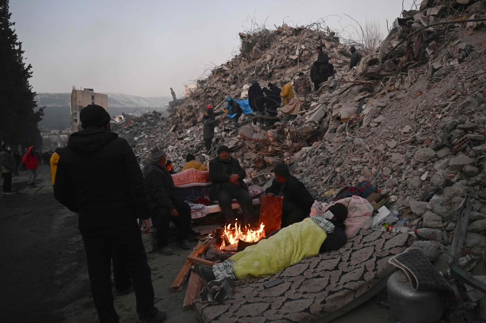 Relatives warm up around a fire in front of rubble of collapsed buildings as rescue teams continue to search victims and survivors, after a 7.8 magnitude earthquake struck the border region of Turkey and Syria earlier in the week, in Kahramanmaras on February 12, 2023. (Photo by Ozan Kose / AFP)
