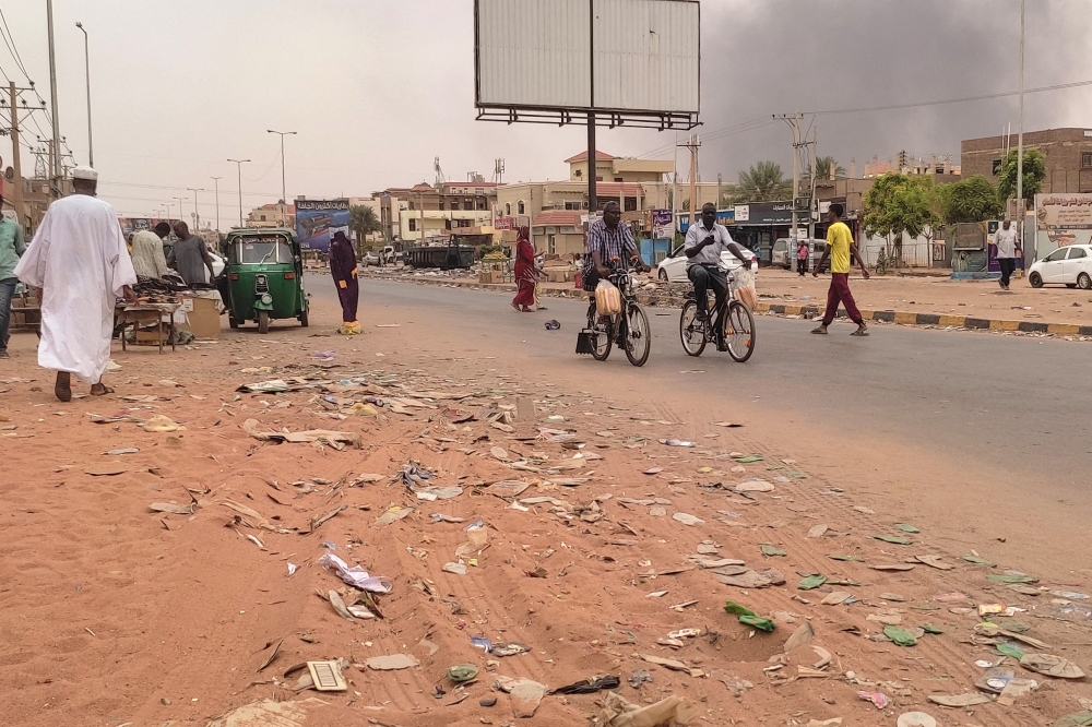 Smoke rises above buildings as people walk along a street in Omdurman on July 15, 2023. Since April 15, 2023, the forces the Sudanese army chief have been at war with the paramilitary Rapid Support Forces (RSF) paramilitary group. (Photo by AFP)