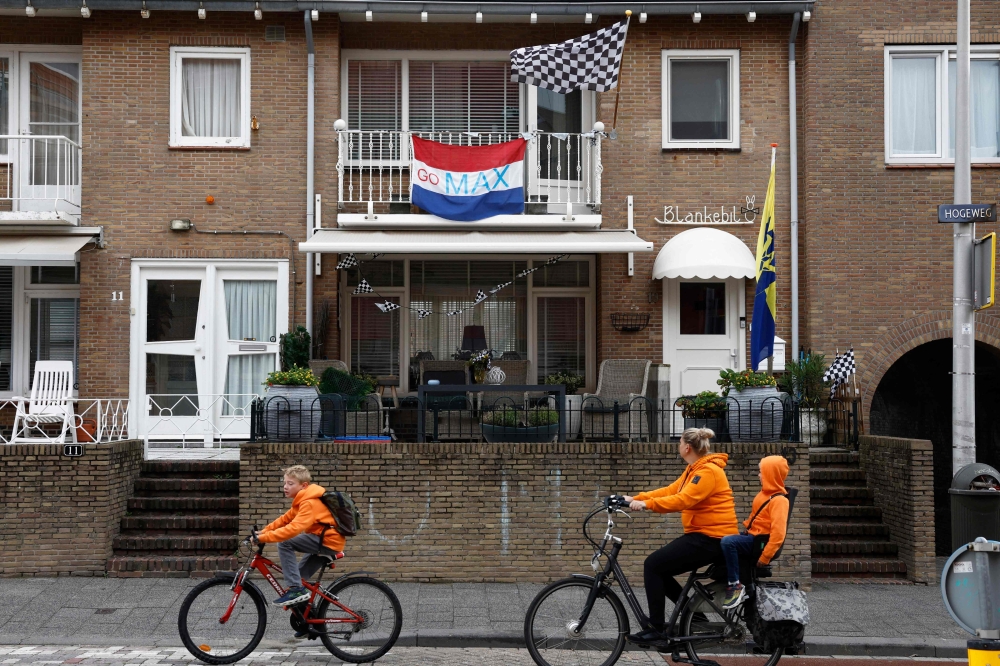 Cyclists dressed in orange ride past a house with a Dutch flag reading