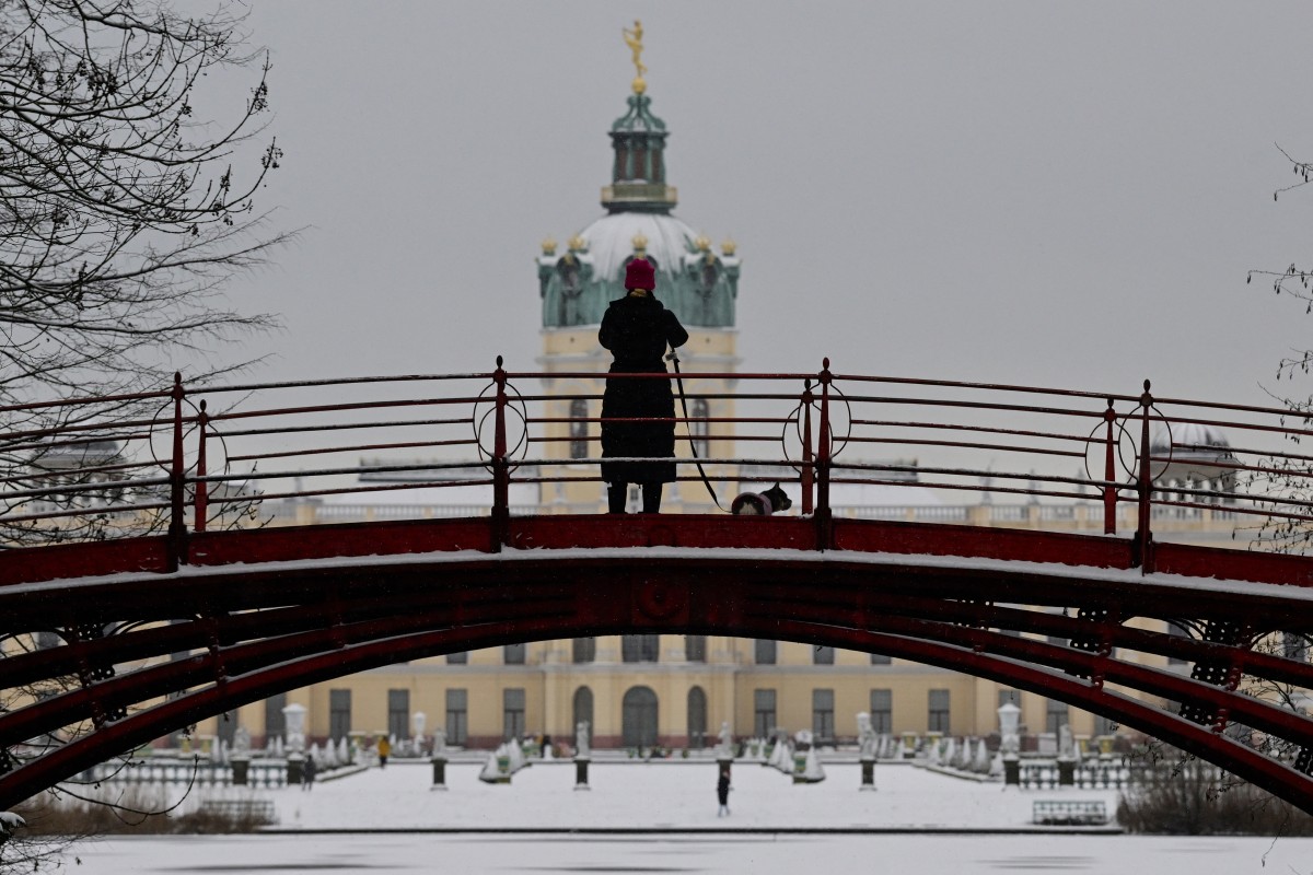 A woman walking her dog, stop to make a photo as she walks across a bridge overlooking the snow-covered gardens of Charlottenburg Palace as snow falls in Berlin on January 16, 2024. (Photo by John MACDOUGALL / AFP)
