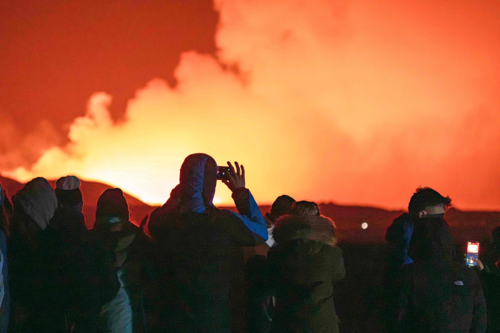 People gather to watch as molten lava flows out from a fissure on the Reykjanes peninsula north of the evacuated town of Grindavik, western Iceland on March 16, 2024 Photo Credit: Ael Kermarec / AFP.