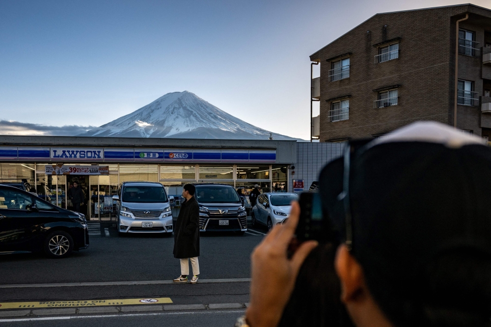 This photo taken on January 1, 2024 shows a tourist posing in front of a convenience store with Mount Fuji in the background, in the town of Fujikawaguchiko, Yamanashi prefecture. Photo by Philip FONG / AFP