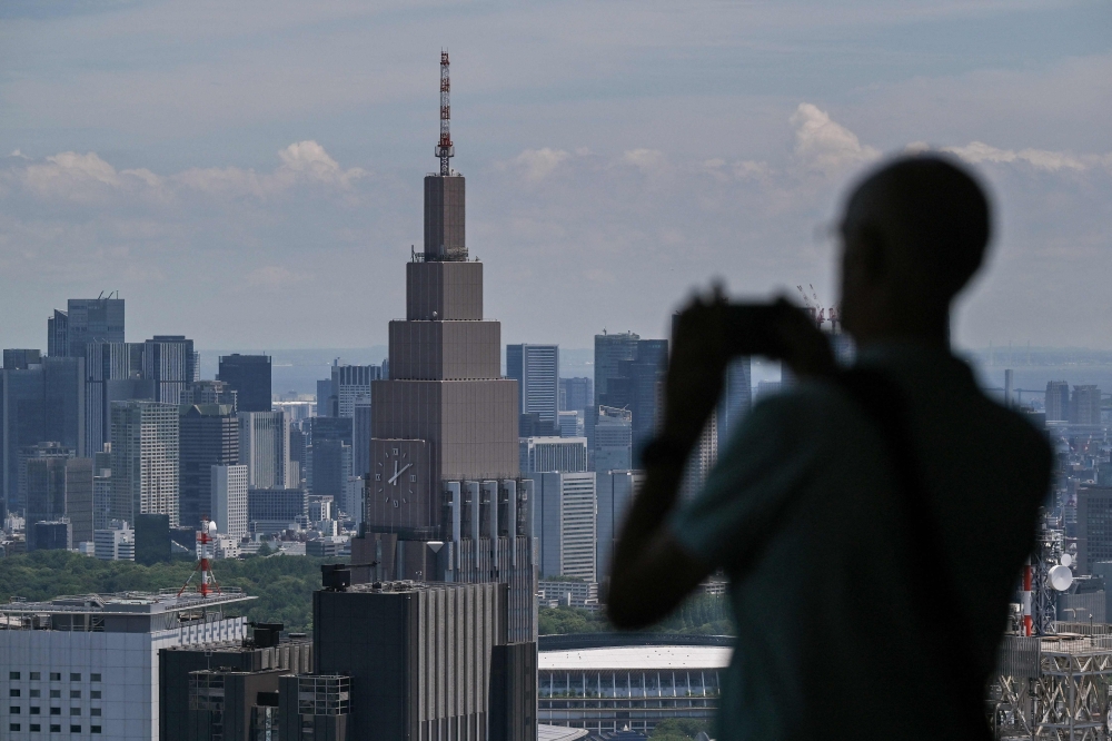 A visitor takes a photo out the window toward the 240m tall (790 ft.) NTT Docomo Yoyogi highrise building (back C) from the viewing deck at the Tokyo Metropolitan Government building in the Shinjuku area of central Tokyo on June 12, 2024. (Photo by Richard A. Brooks / AFP) 