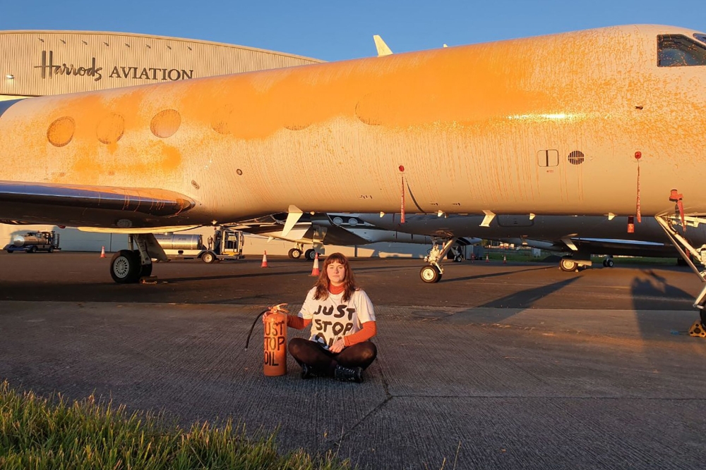 A handout picture released by the Just Stop Oil climate campaign group shows an activist sitting in front of a plane after spraying orange paint on private jets at Stansted Airport, northeast of London, on June 20, 2024. (Photo by Handout / Just Stop Oil / AFP) 
