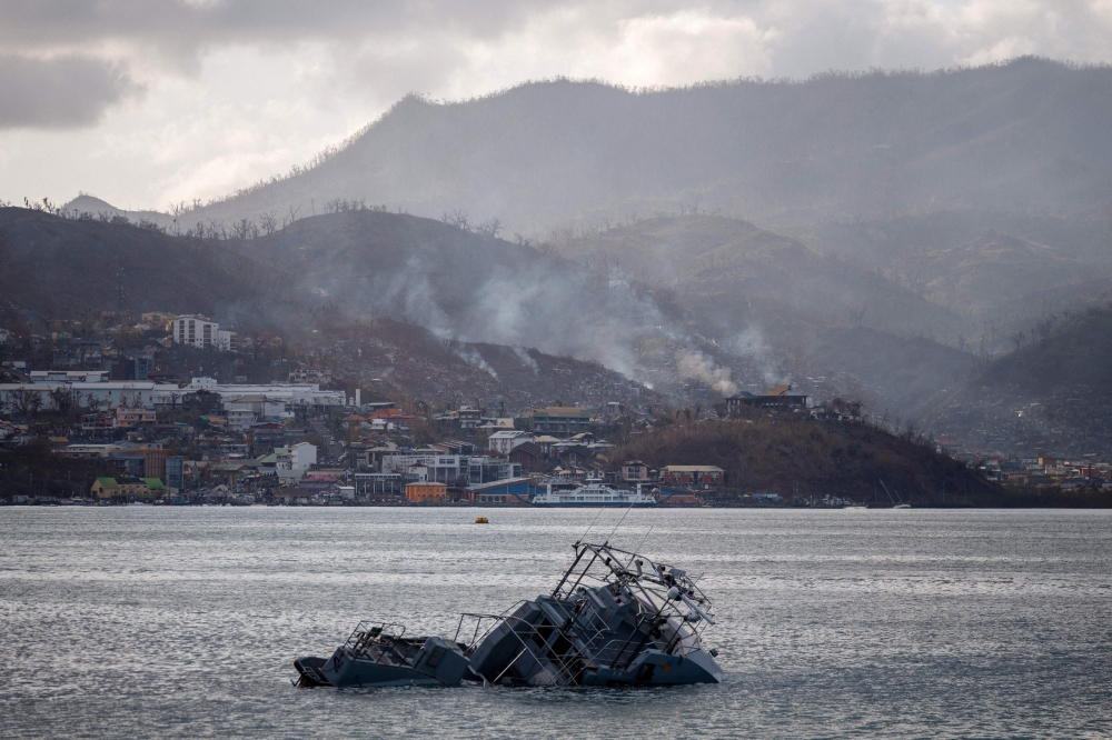 This photograph shows a sinked boat and in background damages and smoke on hills near the city of Mamoudzou, on the French Indian Ocean territory of Mayotte, on December 17, 2024, after the cyclone Chido hit the archipelago. (Photo by Dimitar Dilkoff / AFP)