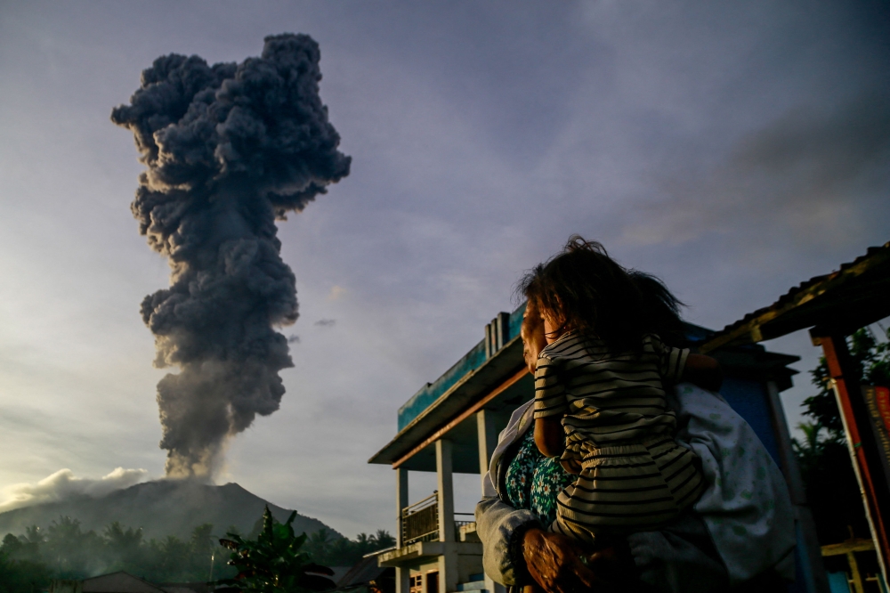 A woman and child look on at volcanic ash rising into the air during the eruption of Mount Ibu, as seen from Duono Village in West Halmahera, North Maluku province, on January 15, 2025. (Photo by Azzam / AFP)
 