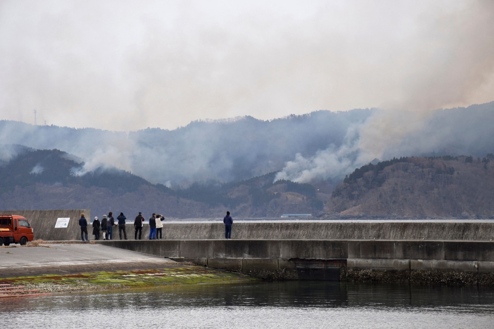 This picture taken on March 3, 2025 shows people watching a wildfire which approaches a village from the fishing port of Ofunato city in Iwate prefecture. (Photo by JIJI PRESS / AFP) 