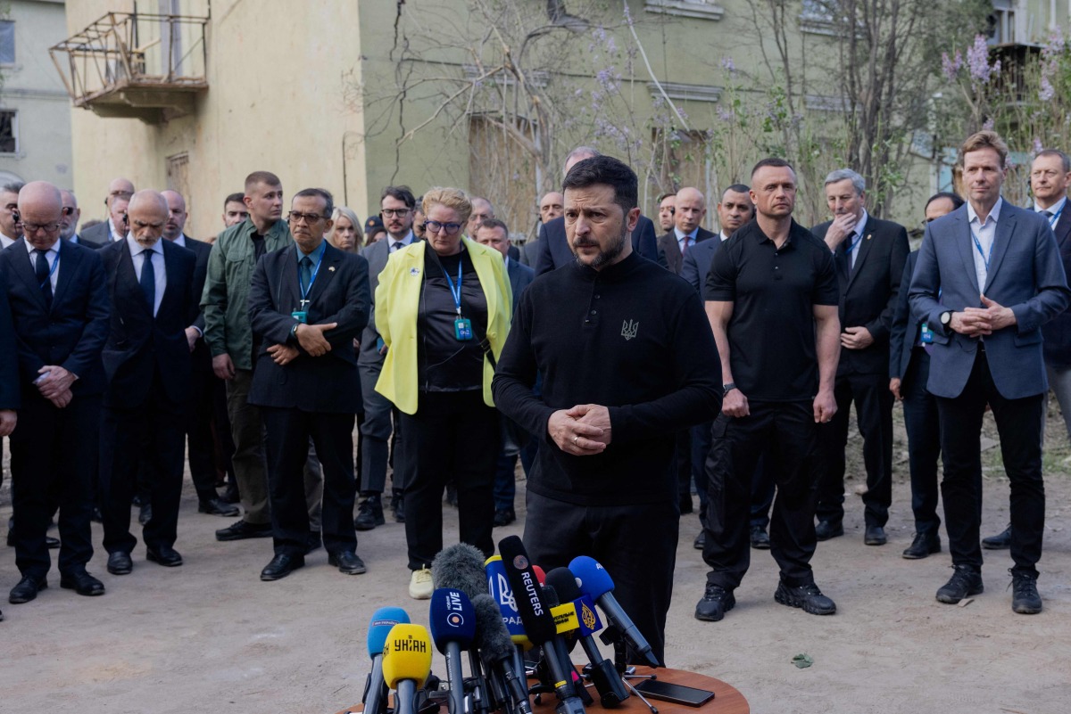 Ukraine's President Volodymyr Zelensky talks to the press as diplomats stands behind him near a destroyed residential building following a Russian missile attack in Kyiv on April 25, 2025, amid the Russian invasion of Ukraine. (Photo by Tetiana DZHAFAROVA / AFP)
