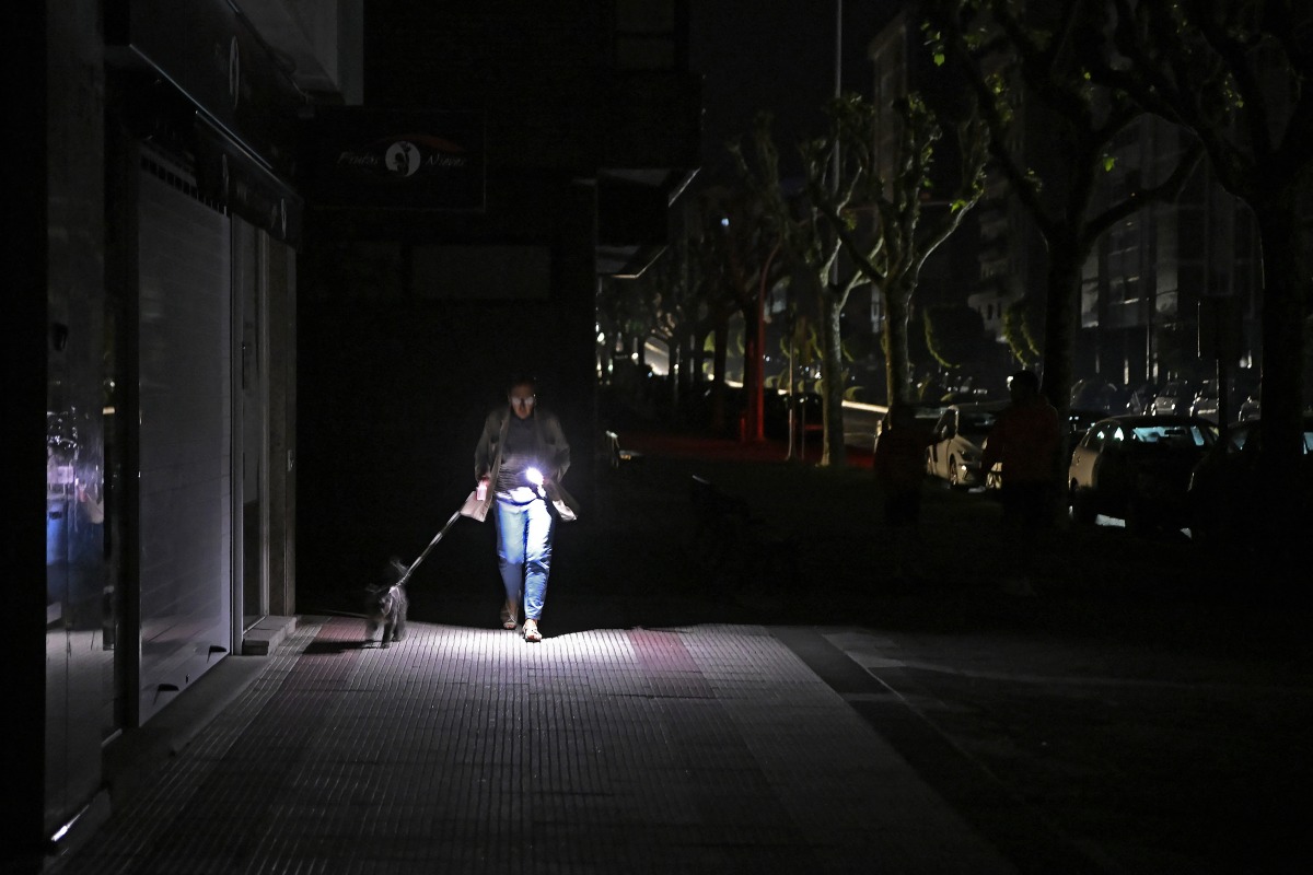 A woman uses her phone's torch while she walks her dog as the street lies in complete darkness during a massive power cut affecting the entire Iberian peninsula and the south of France, in Vigo on April 28, 2025. Photo by MIGUEL RIOPA / AFP