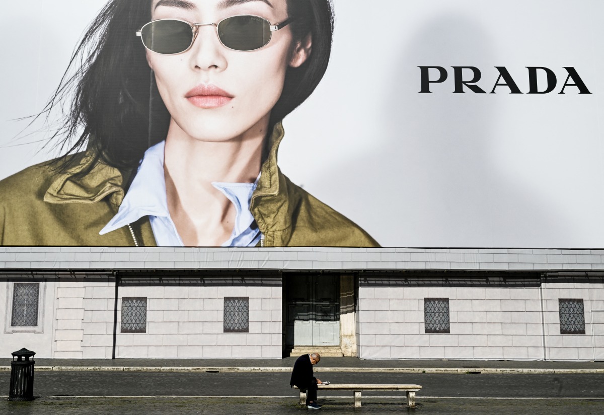 A man sits on a bench reading a newspaper next to a billboard of Italian luxury fashion brand Prada, on Navona Square on April 27, 2025. (Photo by Stefano RELLANDINI / AFP)
