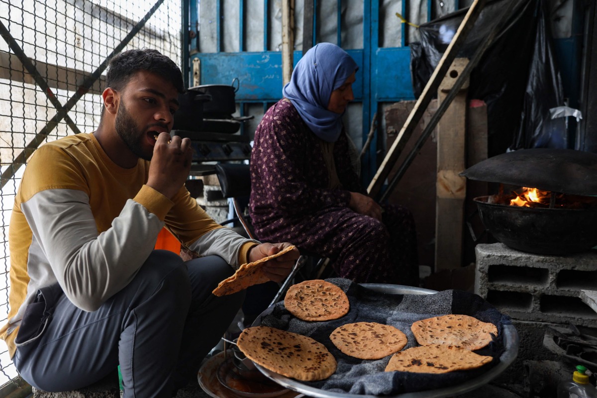 A Palestinian man eats flat bread made with ground lentils during a flour shortage, as woman bakes a new batch in the Jabalia camp for refugees in the northern Gaza Strip on May 6, 2025. Photo by Omar AL-QATTAA / AFP