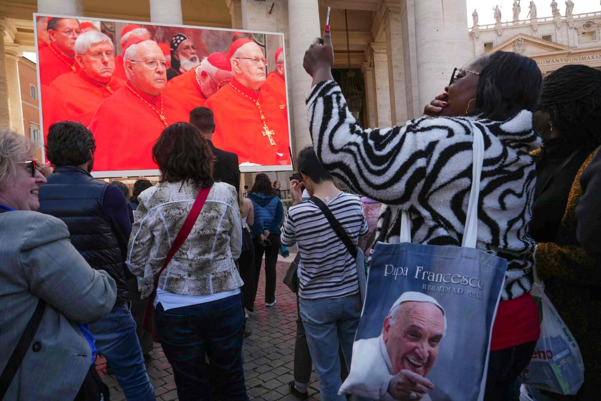 An attendee wearing a bag depicting the late Pope Francis as she watches a giant screen displaying images of the holy mass of cardinals to the Sistine chapel, at St Peter's Square on the first day of the conclave to elect the next pope, at the Vatican, on May 7, 2025. (Photo by Dimitar DILKOFF / AFP)
