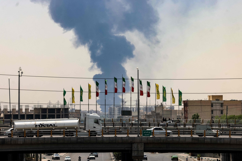 Flags flutter along a bridge as a plume of heavy smoke and fire rise from an oil refinery in southern Tehran, after it was hit in an overnight Israeli strike, on June 15, 2025.  (Photo by Atta Kenare / AFP)
 