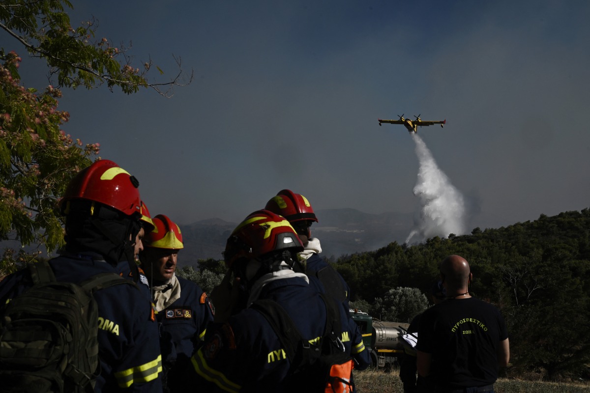 Firefighters gather on a field near the area where an airplane drops water over a wildfire that broke up in Pikermi, some 30 Kms east of Athens on July 3, 2025. Photo by Aris MESSINIS / AFP.
