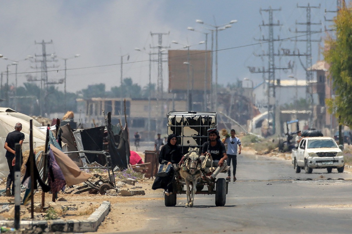 A man and woman ride in a donkey-drawn cart carrying a water cistern while behind smoke billows from Israeli bombardment, along Salah al-din road in Deir el-Balah in the central Gaza Strip on July 21, 2025. (Photo by Eyad BABA / AFP)