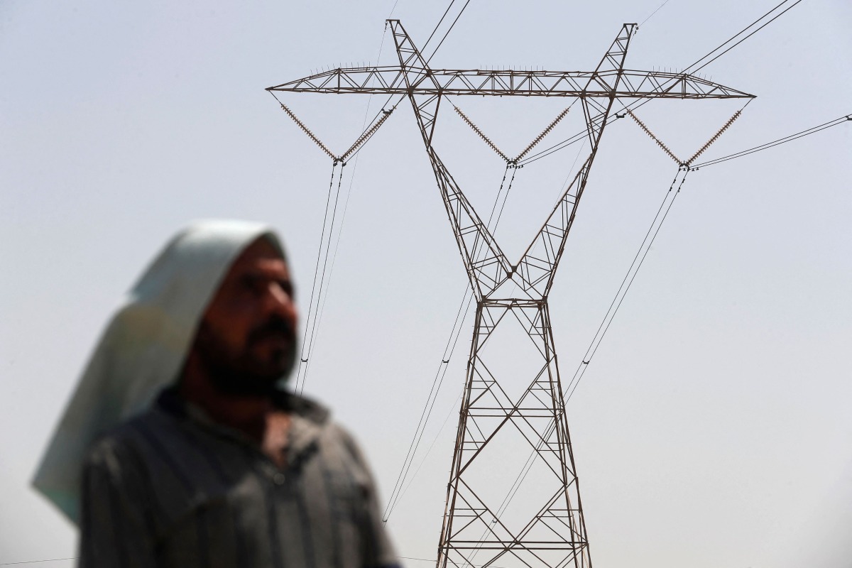 An Iraqi man stands under an electricity transmission tower south of Hilla city on August 10, 2025, amidst rising temperatures, chronic water shortages and power cuts. (Photo by AHMAD AL-RUBAYE / AFP)
