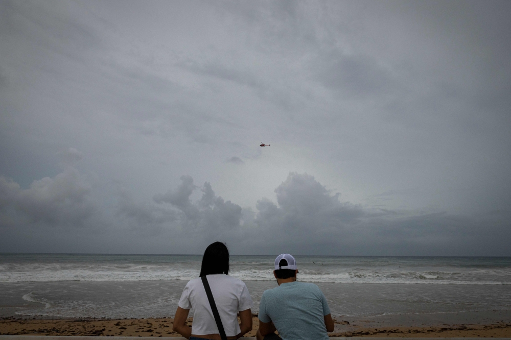 A helicopter flies over as a couple looks at La Pared beach as Category 5 Hurricane Erin approaches in Luquillo, Puerto Rico on August 16, 2025. (Photo by Ricardo Arduengo / AFP)