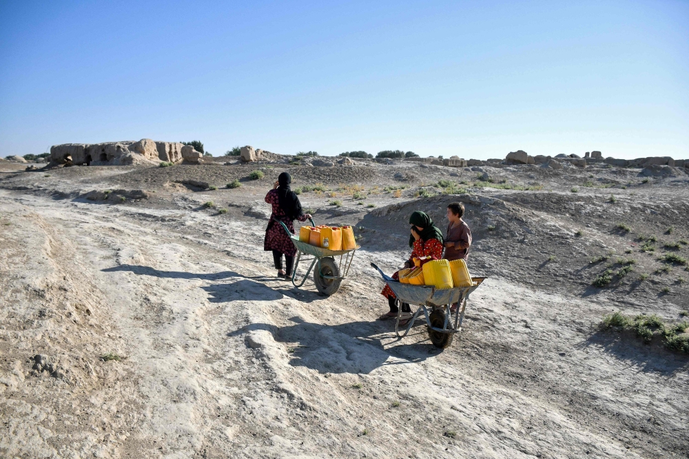 An Afghan woman with children takes a break as they haul water cans on wheelbarrows along a deserted street in the drought-ridden village of Bolak at Chahar Bolak district, Balkh province on July 10, 2025.  (Photo by Atif Aryan / AFP)
