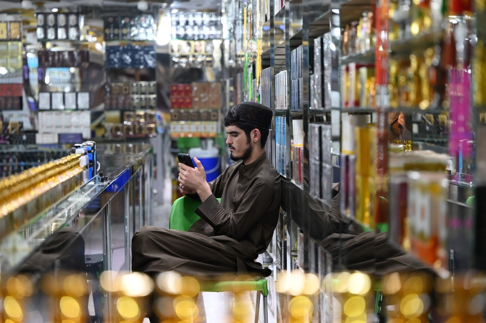An Afghan vendor selling perfume and body sprays, uses his mobile phone as he sits at his shop in Kabul on October 1, 2025.  (Photo by Wakil Kohsar / AFP)
