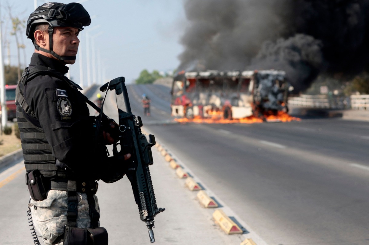 A member of the Prosecutor's Office stands guard near a burning bus at one of the main avenues after it was set on fire by organised crime groups in response to an operation in Jalisco to arrest a high-priority security target in Zapopan, state of Jalisco, Mexico, on February 22, 2026. (Photo by Ulises Ruiz / AFP)
