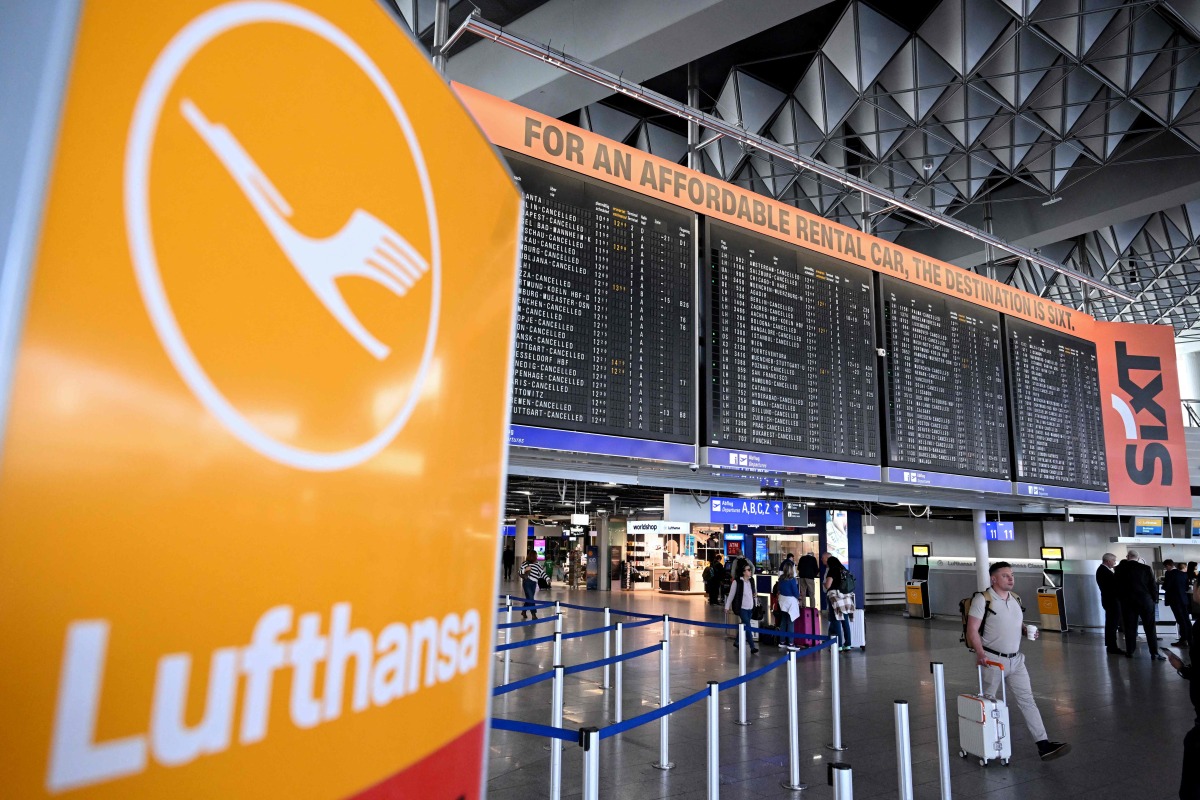 The logo of German airline Lufthansa is seen in the foreground as passengers walk below a flight information display at Frankfurt Airport, Frankfurt am Main, western Germany, on April 15, 2026, as cabin crew union UFO urged members of Lufthansa cabin crew to stage a strike in a dispute over pay and pensions. Photo by Kirill KUDRYAVTSEV / AFP